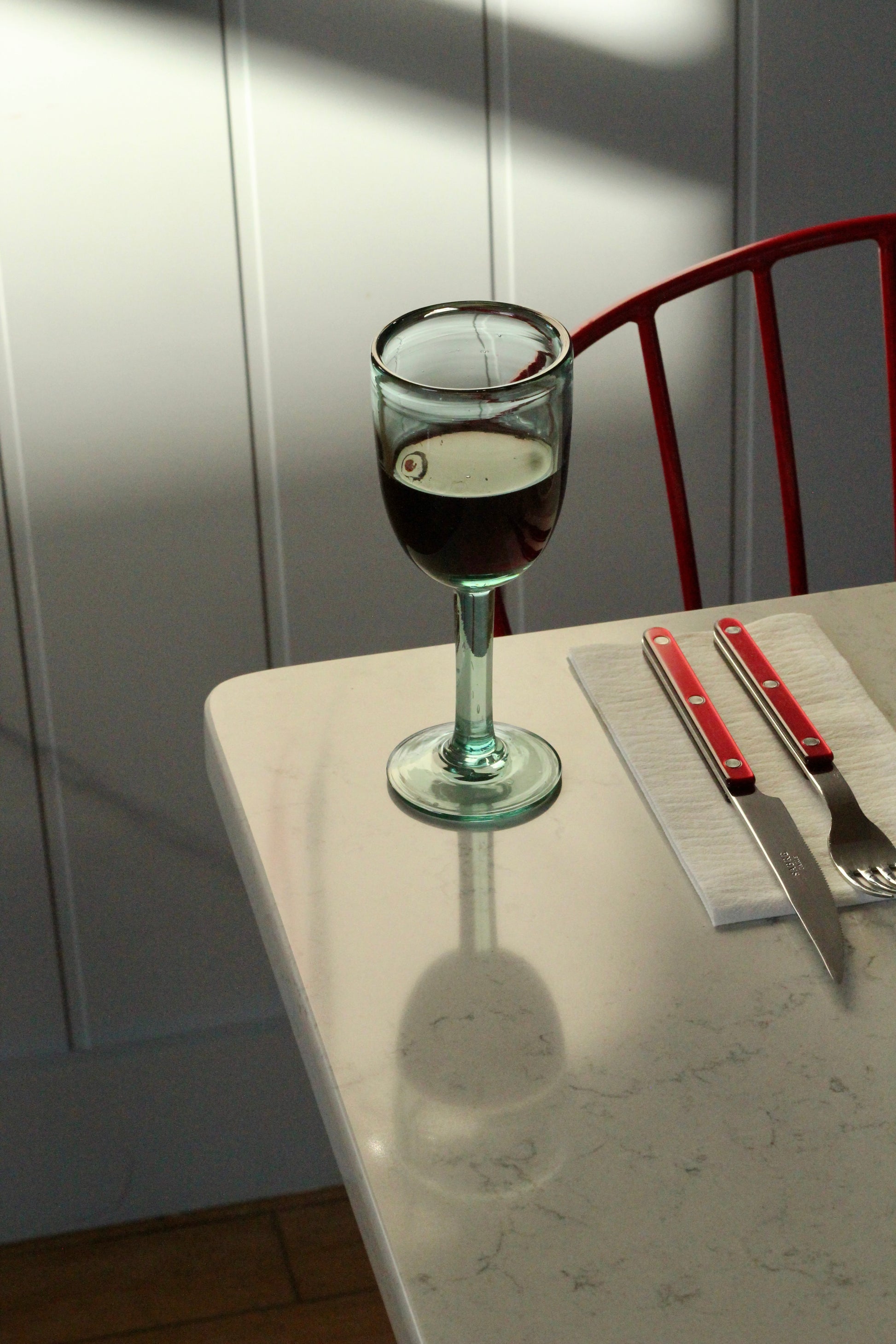 Handblown recycled glass of red wine on a table with cutlery and a red chair in the background