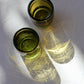 Two handblown mini glasses filled with a clear liquid on a textured white surface.