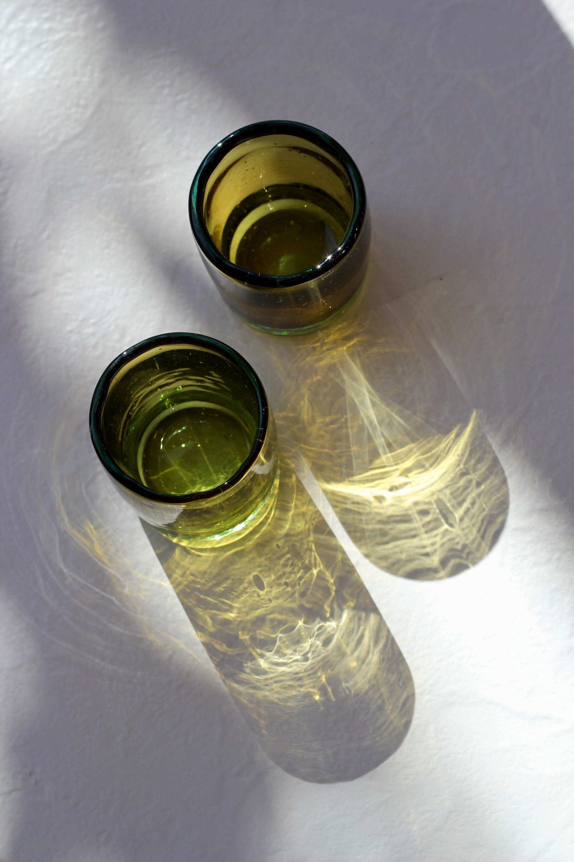 Two handblown mini glasses filled with a clear liquid on a textured white surface.