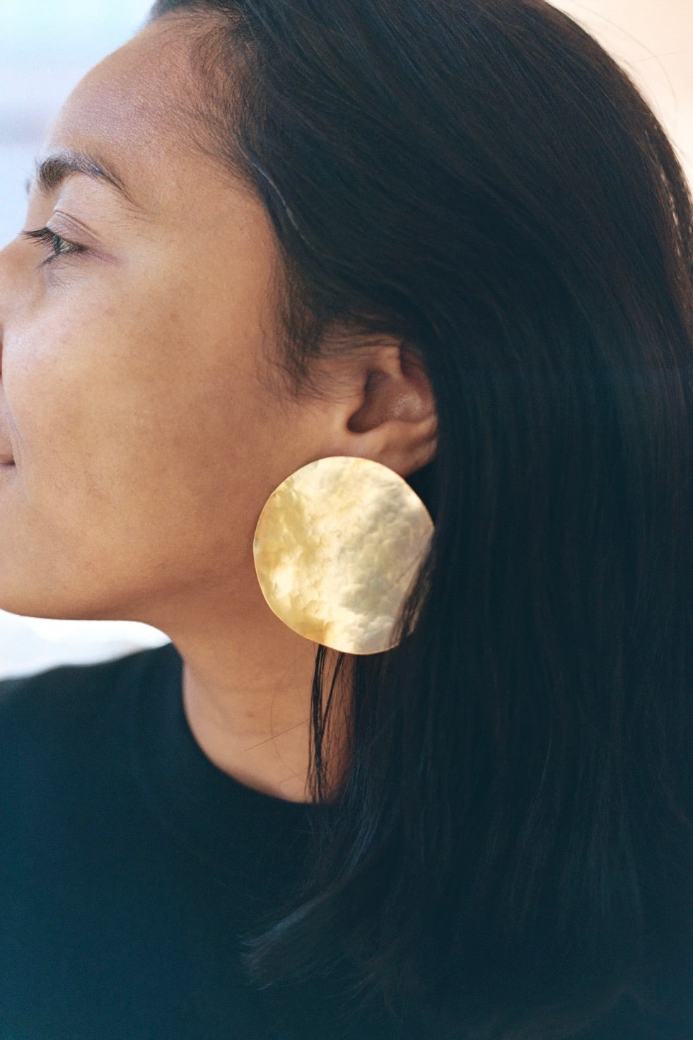 Close-up of a person wearing a large gold hand-hammered earring with a neutral background