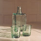 Two green glass tumblers and a handblown glass carafe on a reflective surface with a blurred background