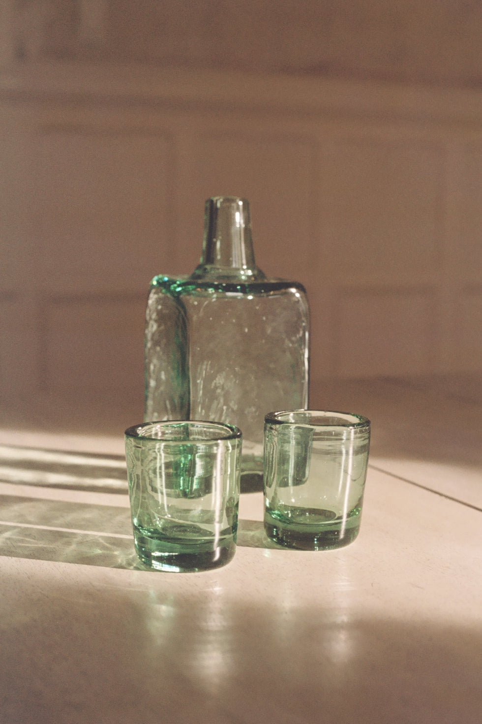 Two green glass tumblers and a handblown glass carafe on a reflective surface with a blurred background