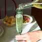 Person pouring champagne into a  handblown glass on a table with food and a chair in the background.