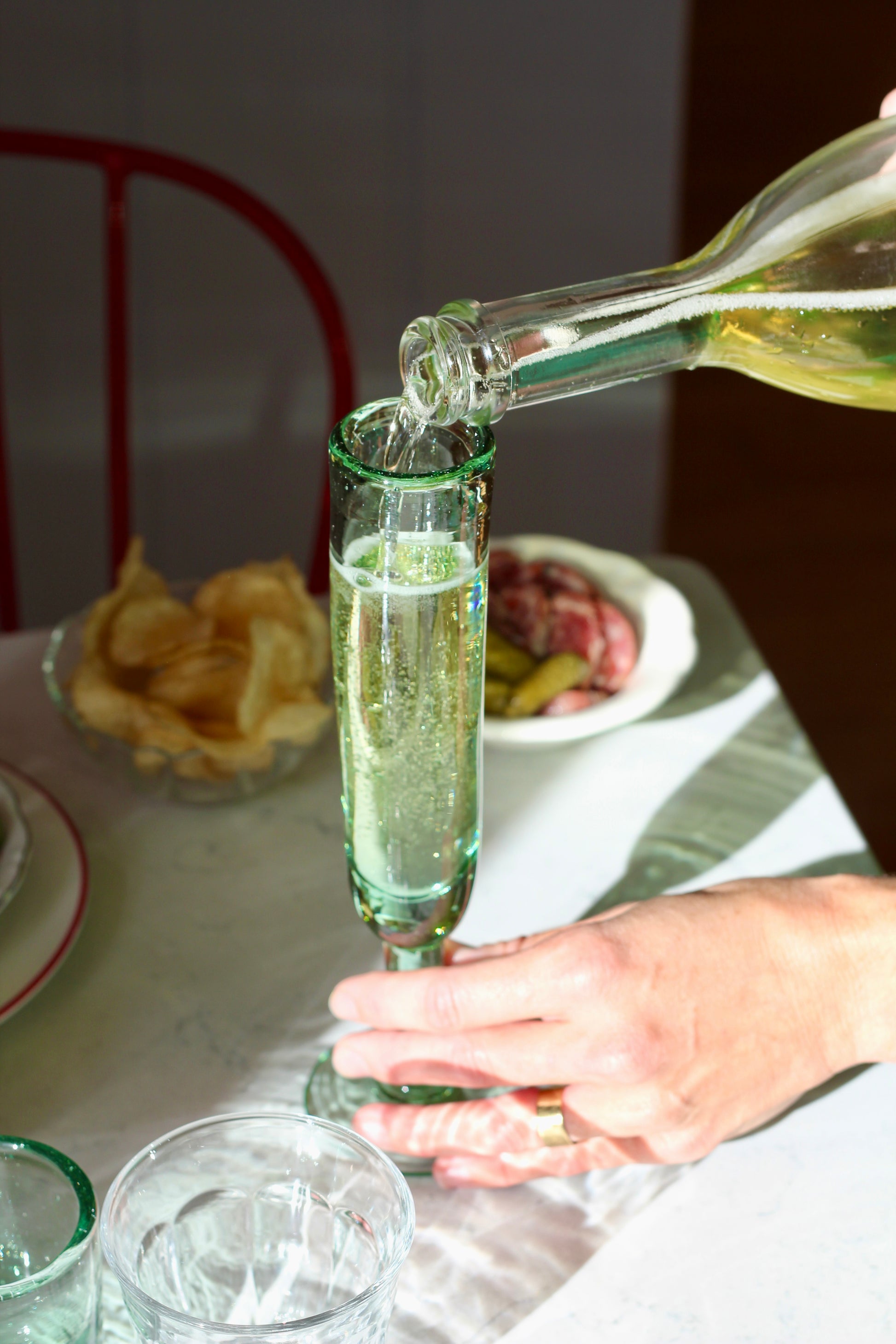 Person pouring champagne into a  handblown glass on a table with food and a chair in the background.