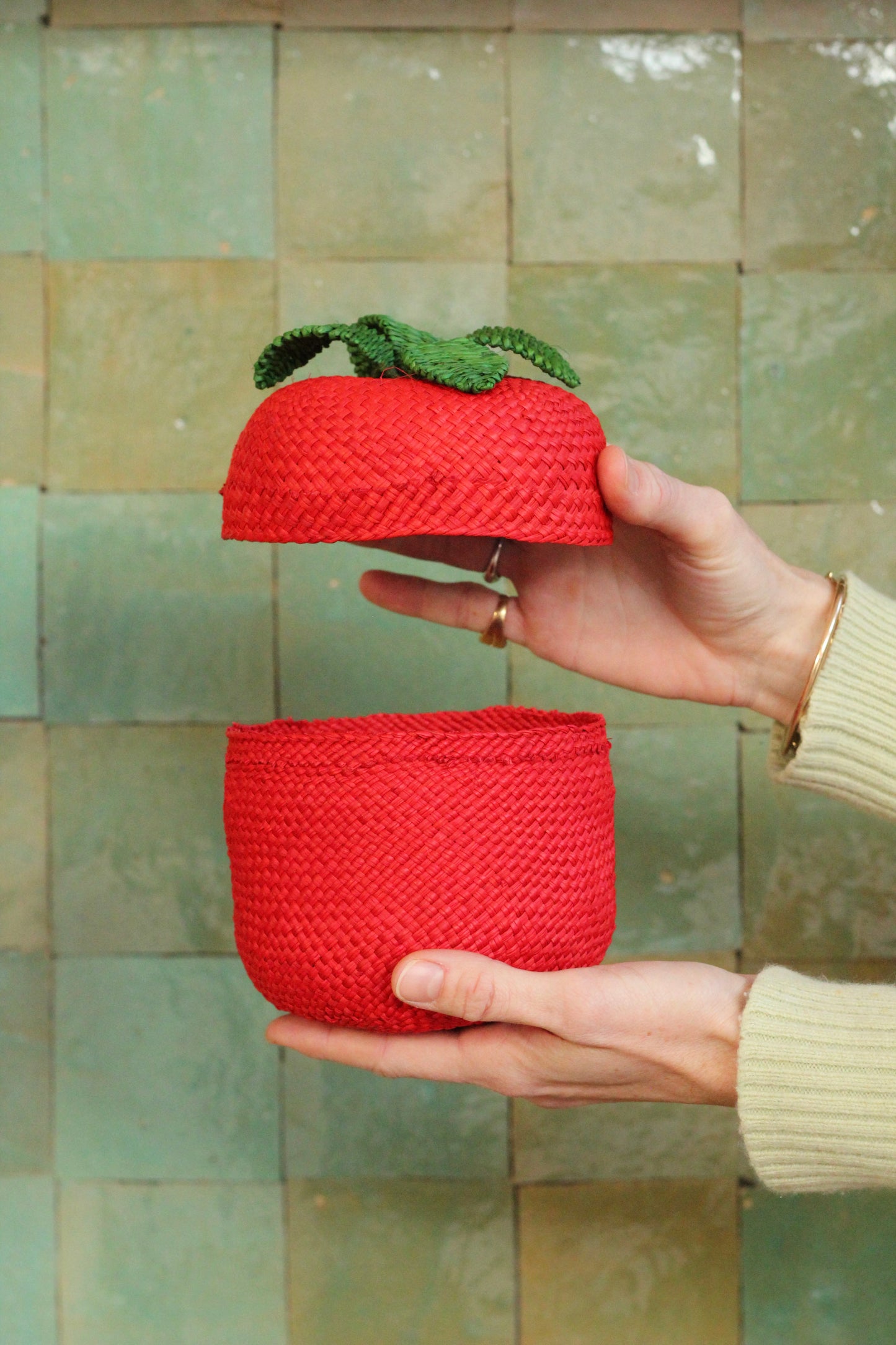 Red woven tomato basket held by a hand against a tiled wall.