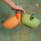 Hand holding two mini woven fruit baskets, one orange and one green, against a tiled wall.