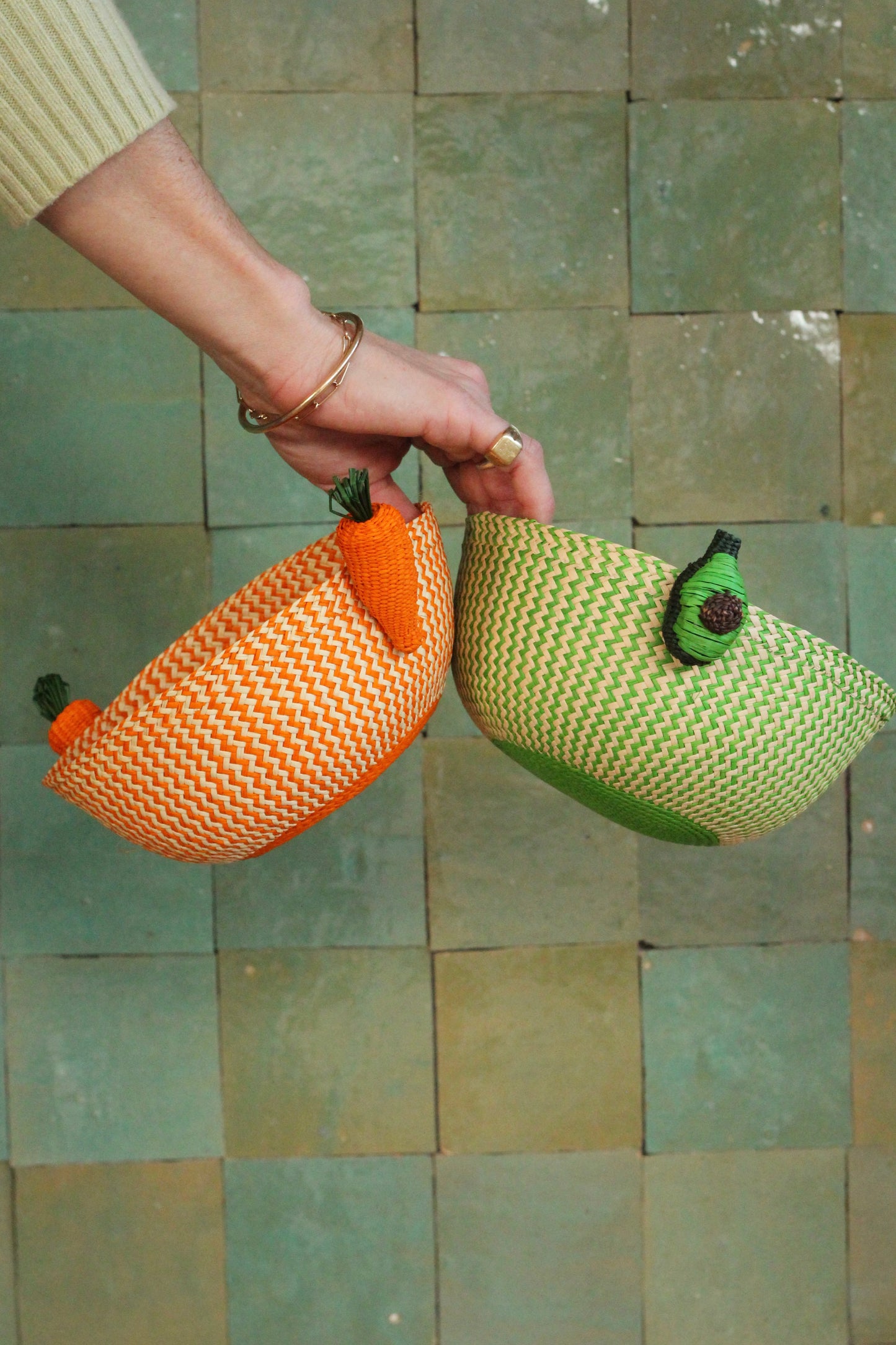 Hand holding two mini woven fruit baskets, one orange and one green, against a tiled wall.