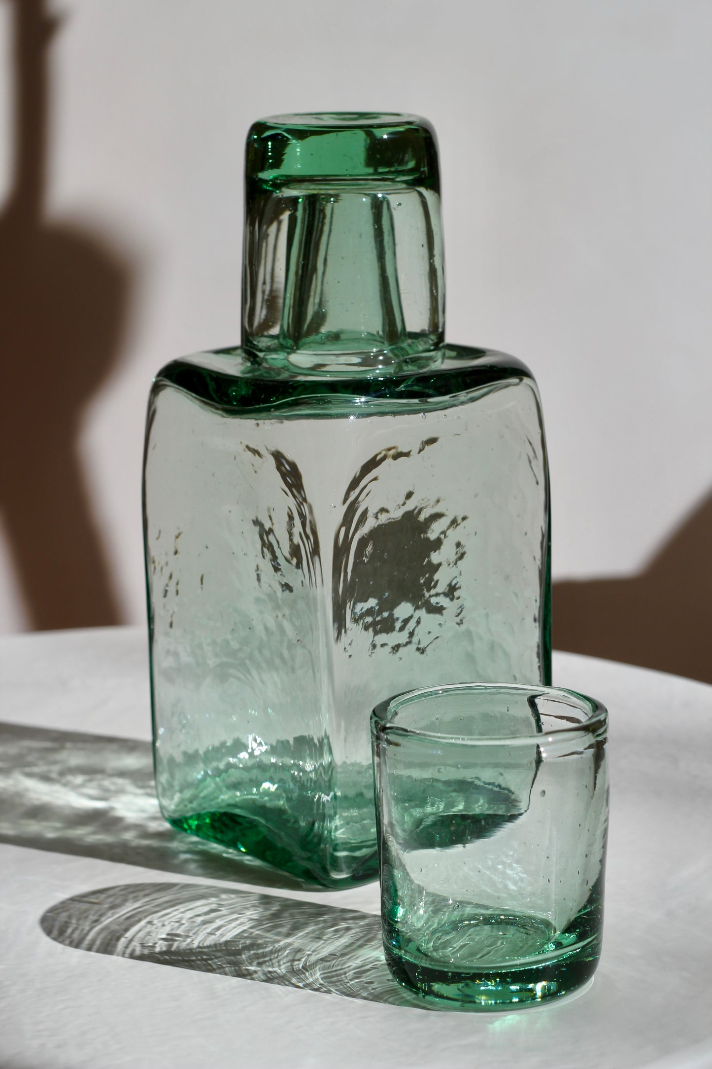Green handblown recycled glass carafe and glass on a white surface with shadows.