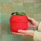 Hand holding a red woven tomato basket with green top against a tiled wall.