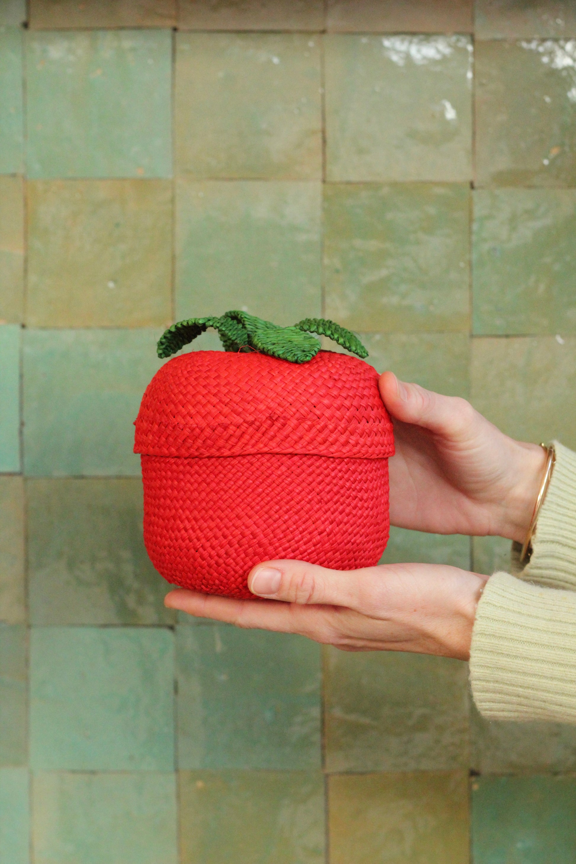 Hand holding a red woven tomato basket with green top against a tiled wall.