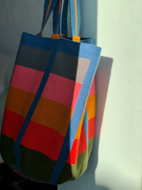 An oversized handwoven tote bag with multicolored stripes in red, blue, green, and yellow, with a black base and colorful straps.